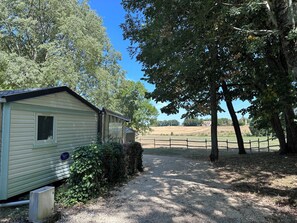 A single-story mobile home with a small window, surrounded by trees and a gravel path. - 2-Bedroom Family Mobile Home – Peaceful & Natural Setting in Castillonnès (Castillonnès)