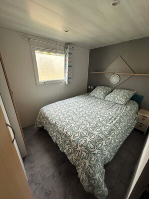 A bedroom with a bed, a window with curtains, a bedside table, and a wall-mounted light fixture. - 2-Bedroom Family Mobile Home – Peaceful & Natural Setting in Castillonnès (Castillonnès)