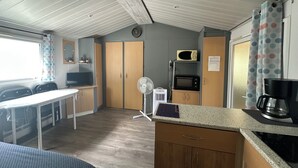 En kompakt tekøkken med mikrobølgeovn, et lille køleskab, kaffemaskine og ventilator. Der er et spiseområde med stole og bord, samt et vindue med gardiner.