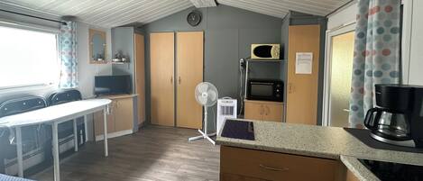 Una kitchenette compacta con microondas, un refrigerador pequeño, una cafetera y un ventilador. Hay una zona de comedor con sillas y una mesa, además de una ventana con cortinas.