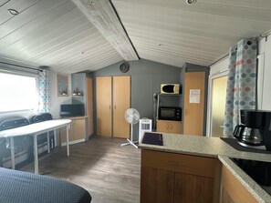 A compact kitchenette with a microwave, a small refrigerator, a coffee maker, and a fan. There is a dining area with chairs and a table, and a window with curtains. - 2-Bedroom Family Mobile Home – Peaceful & Natural Setting in Castillonnès (Castillonnès)