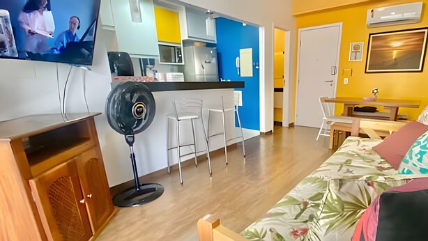 En moderne stue med en træsofa, en gulvventilator og et køkkenområde med en blå accentvæg.