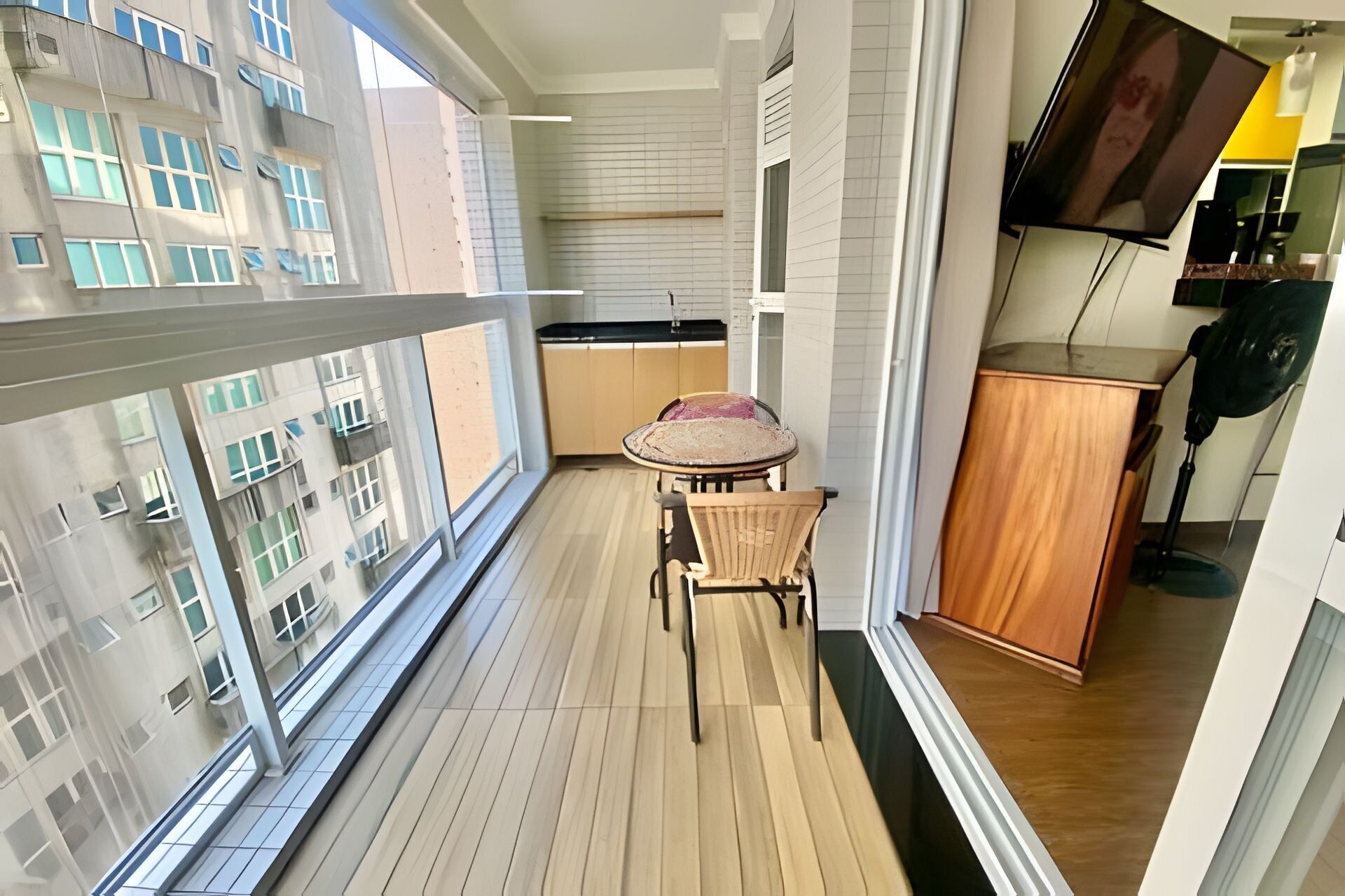 A balcony with a wooden floor, a small table with a chair, a cabinet, and a TV mounted on the wall.