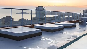 En tagterrasse med gelænder af glas, der viser udsigt over byens skyline og havet i solnedgang.