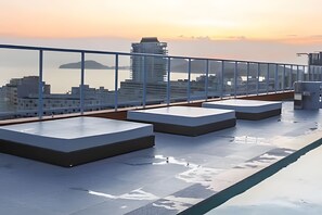 Une terrasse sur le toit, dotée d’une rambarde en verre, qui offre une vue sur le panorama urbain et la mer au coucher du soleil.