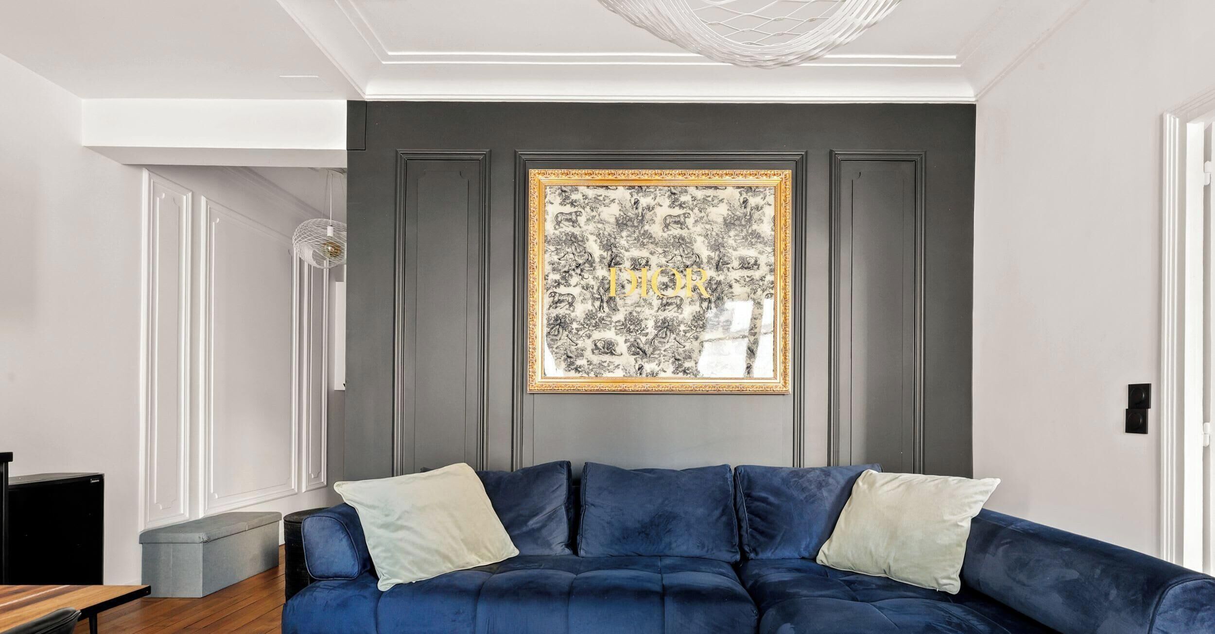 Ein Zimmer mit einem dunkelblauen Sofa, zwei hellen Kissen, einem gerahmten Kunstwerk an der Wand und einer weißen Decke mit einer Leuchte.