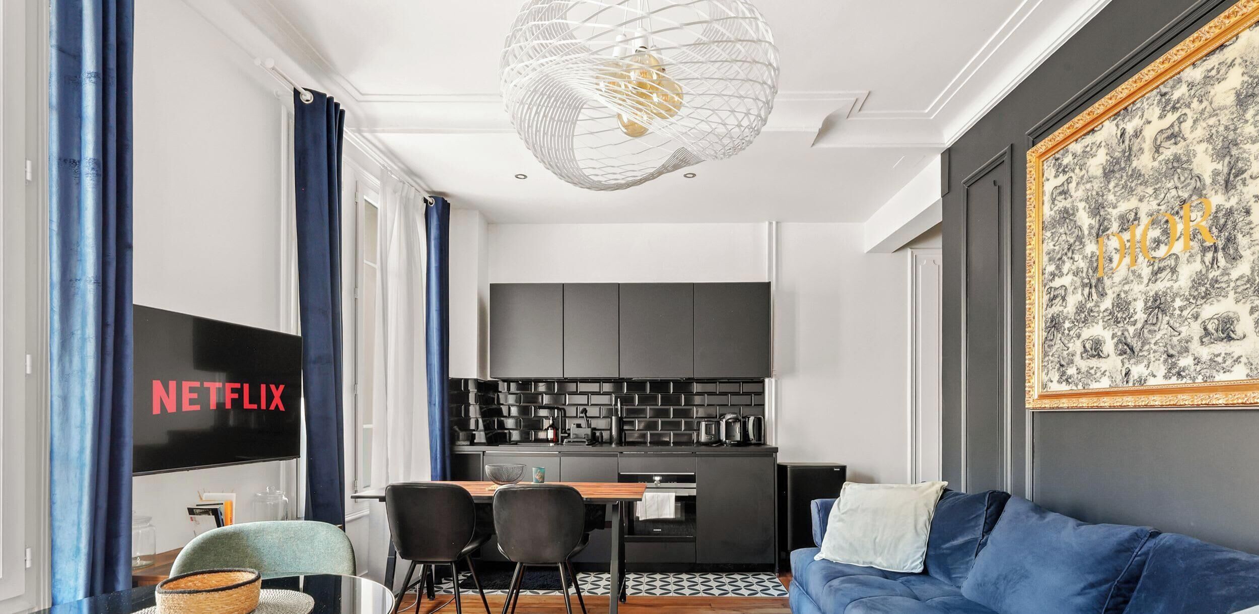 A modern living room with a blue sofa, a dining table, and a kitchen area with grey cabinets.