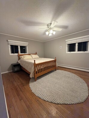 A bedroom with a wooden bed, a round rug, a ceiling fan, and two windows with blinds. - Riverfront escape at Q’s River Place, a 3 bedroom year round getaway (Hersey)