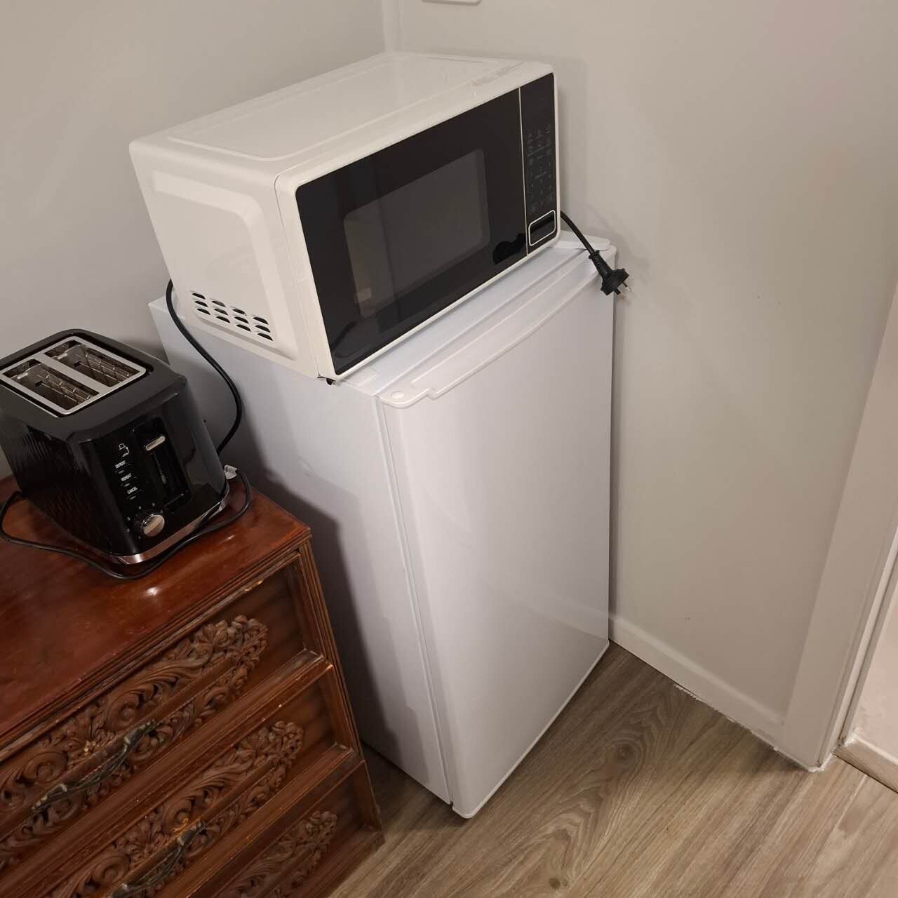 A compact kitchenette with a white microwave on top of a white refrigerator, a black toaster on a wooden sideboard, and a wooden cabinet with intricate carvings.
