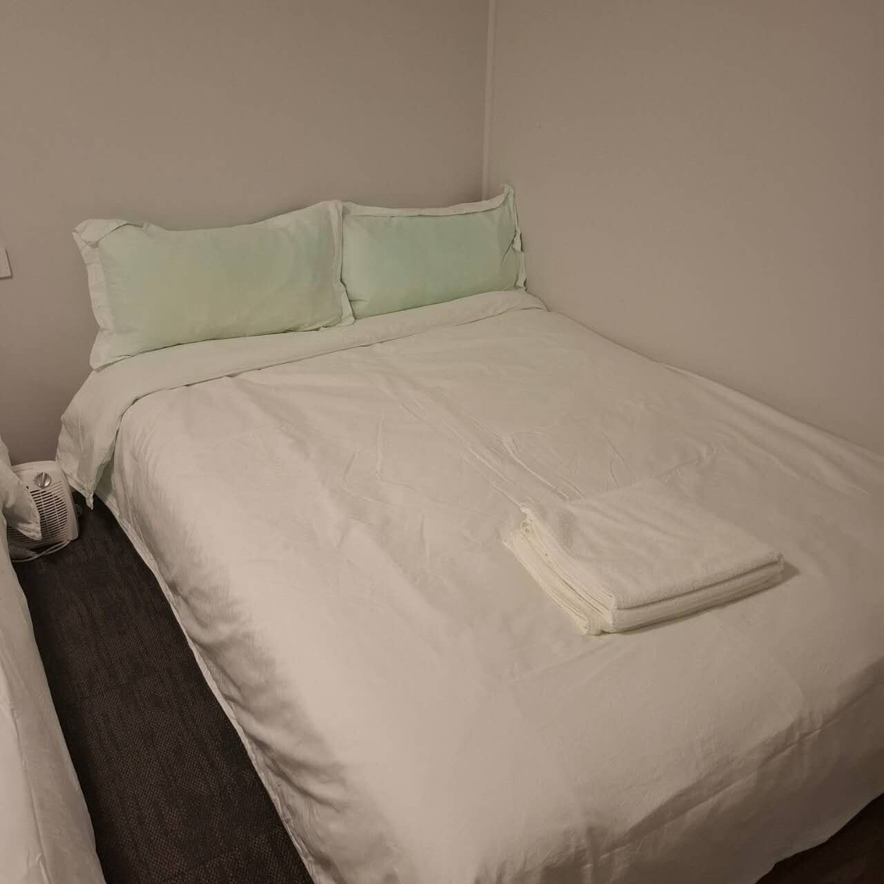 Un lit avec une couette et des oreillers blancs, une serviette pliée posée sur le lit et un petit radiateur sur le sol.