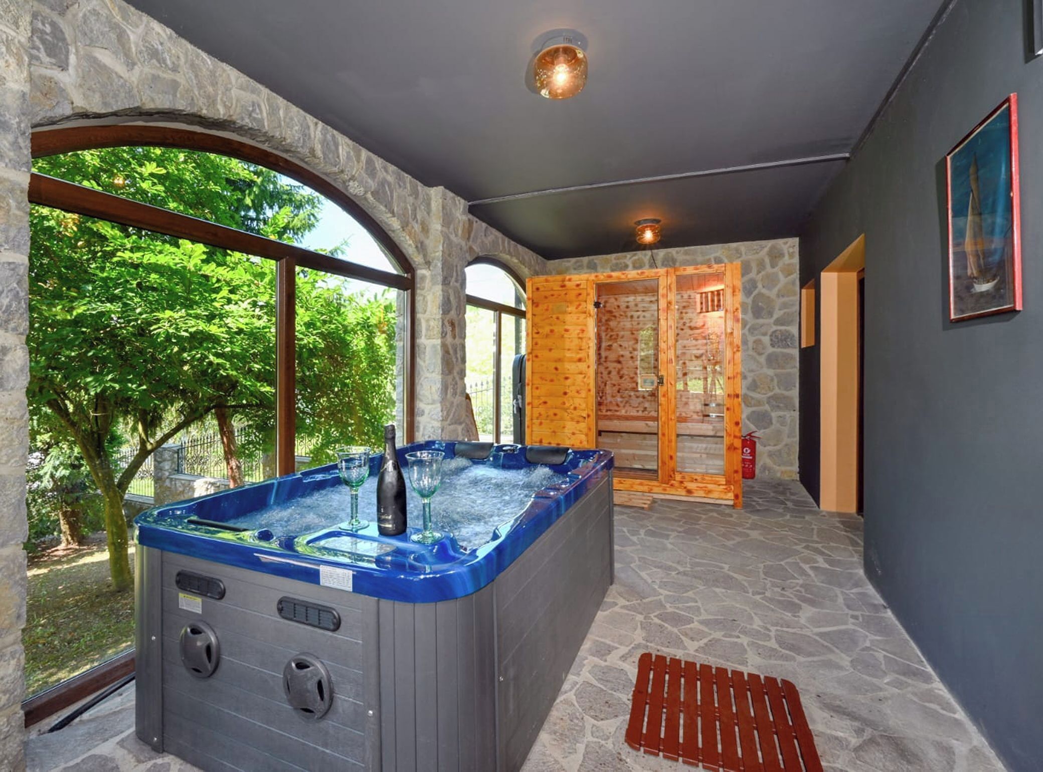 Een wellnessruimte met een hottub, stenen muren en grote ramen met uitzicht op groen.