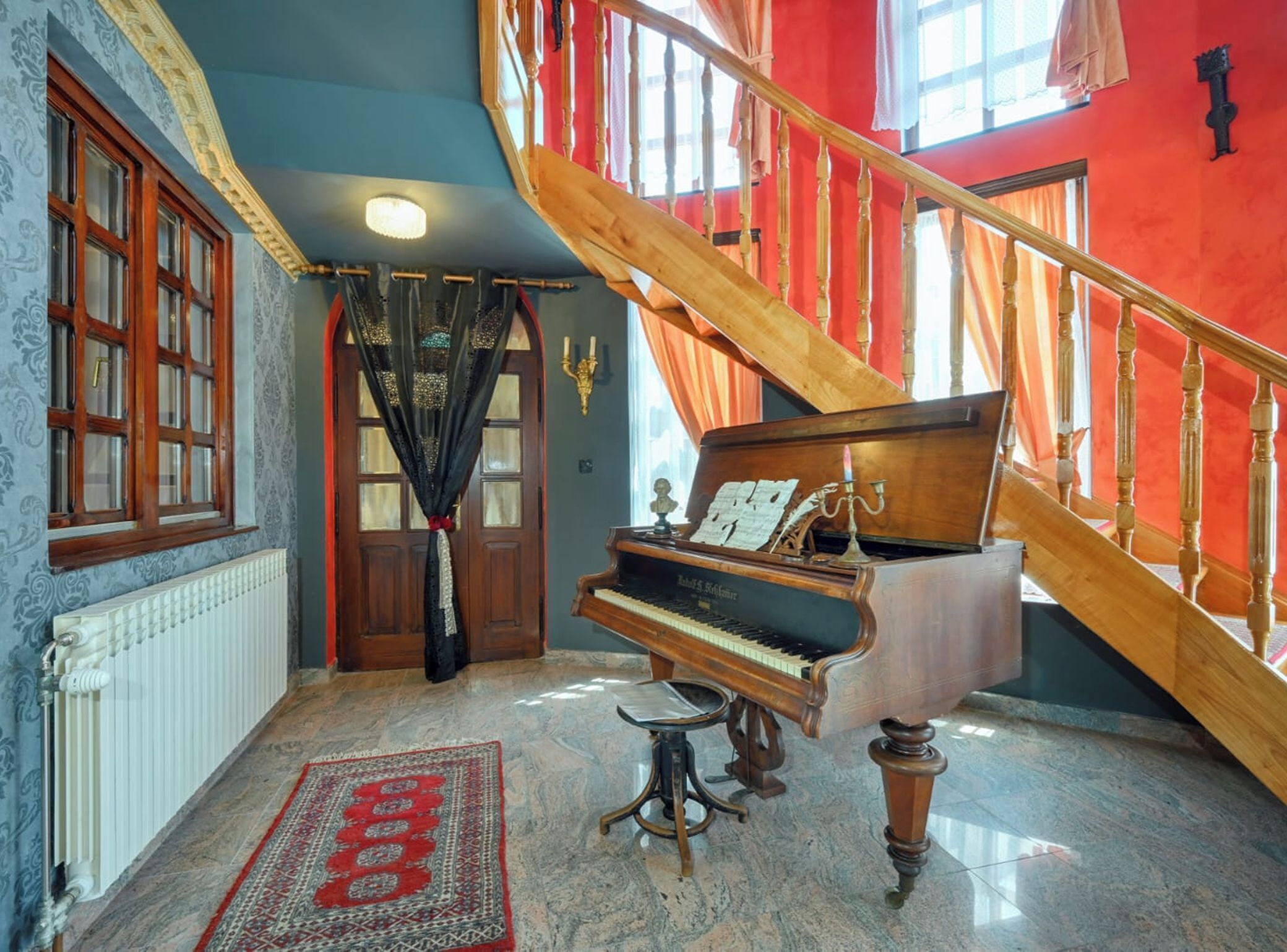 A grand piano in a room with a red staircase, a patterned rug, and a large window with curtains.