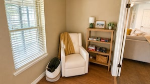 A cozy living room with a beige armchair, a small bookshelf, and a window with blinds.