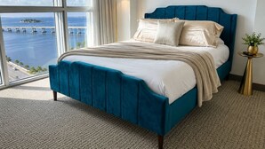 Una cama con cabecera azul, ropa de cama blanca y vista al mar a través de amplios ventanales.