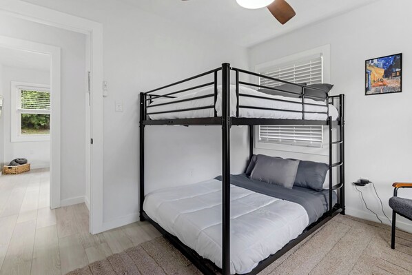 Una cama litera con colchón blanco, manta gris y almohada gris. Se observa una ventana con persianas, un cuadro en la pared y una silla con reposabrazos de madera.
