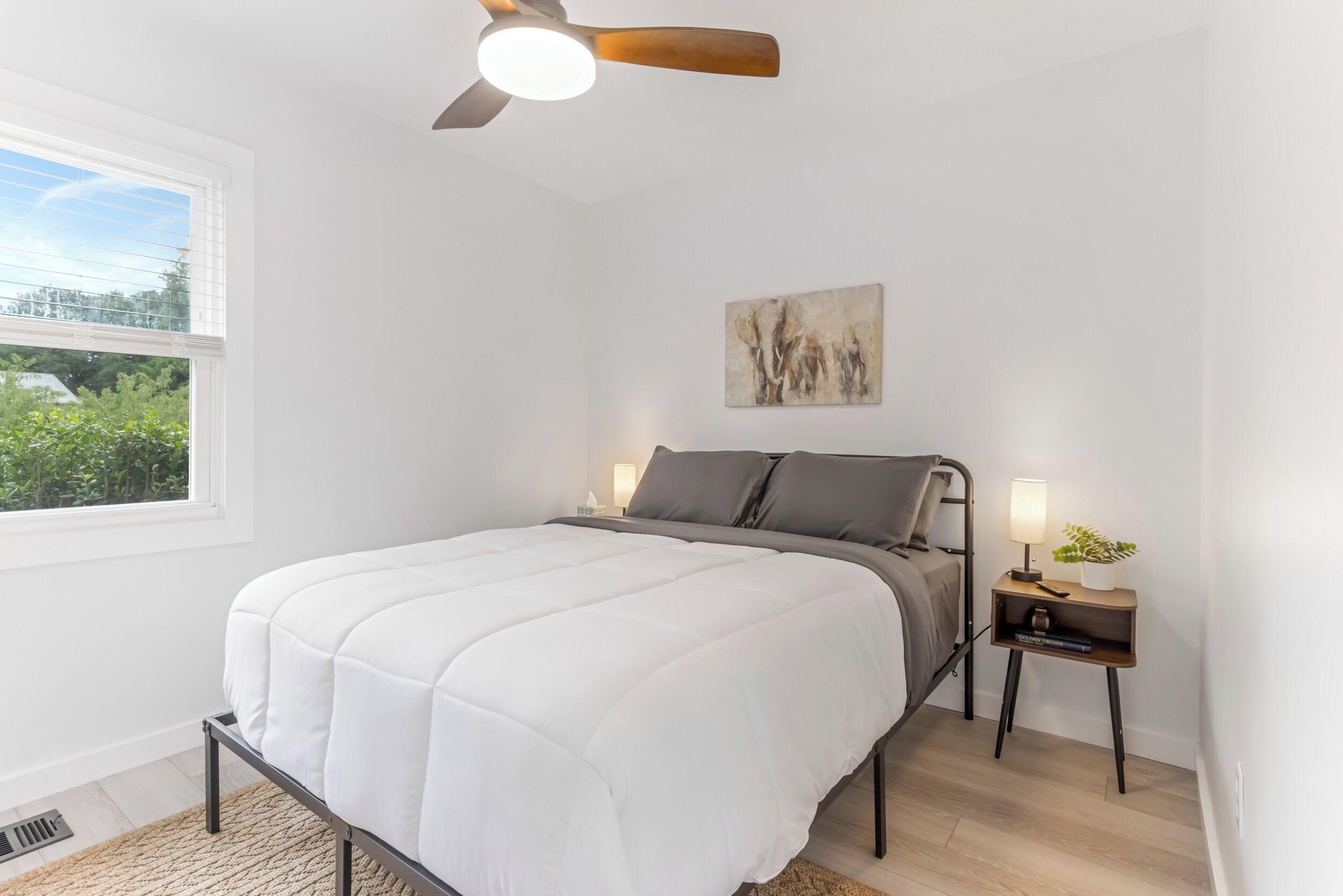 A bedroom with a bed, a ceiling fan, a bedside table with a lamp, and a window with a view of greenery.