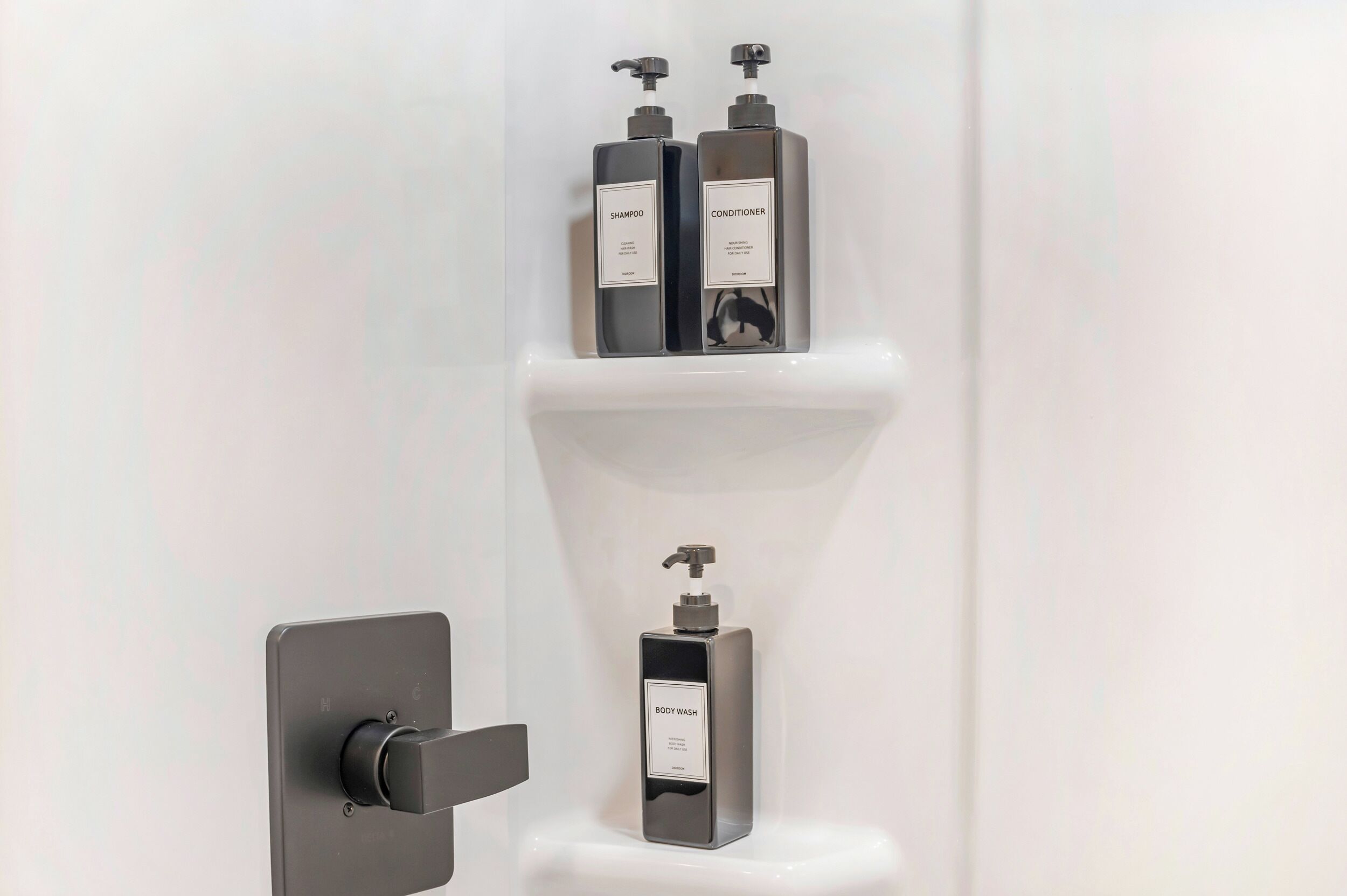 Une salle de bain avec un robinet moderne, une étagère sur laquelle se trouvent deux bouteilles étiquetées « SHAMPOO » et « APRÈS-SHAMPOO », et une seule bouteille étiquetée « GEL DOUCHE ».