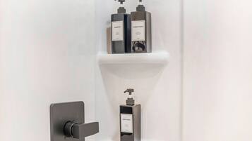 Une salle de bain avec un robinet moderne, une étagère sur laquelle se trouvent deux bouteilles étiquetées « SHAMPOO » et « APRÈS-SHAMPOO », et une seule bouteille étiquetée « GEL DOUCHE ».