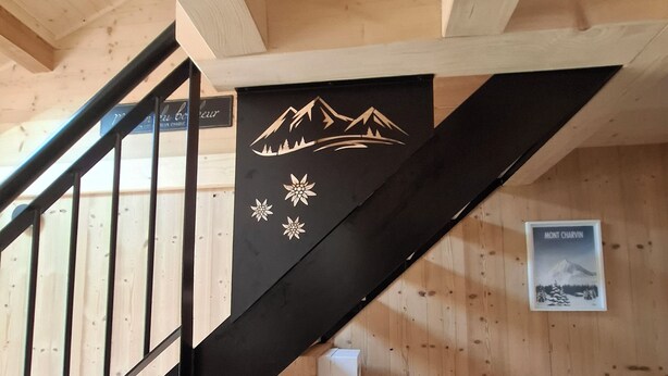 Un escalier en bois avec une rampe noire, un lit recouvert d’une couverture blanche et un mur orné d’un tableau encadré et d’une banderole noire décorée de motifs représentant des montagnes et des fleurs.