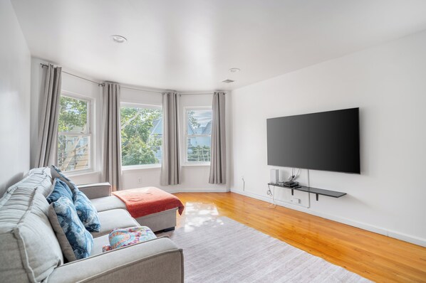A modern living room with a flat-screen TV mounted on the wall, a sofa with cushions, a coffee table, and a large window with curtains. - Cozy 3-bedroom apartment in fantastic Jersey City, easy access to NYC (Jersey City)