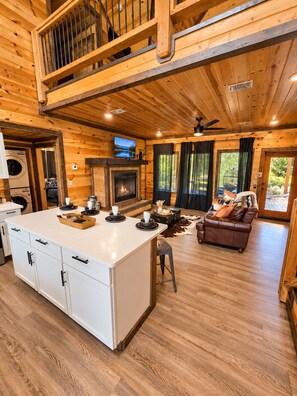 Ein Ferienhaus-Innenraum mit einer zentralen Kochinsel, Kamin, Fernseher und einer gemütlichen Sitzecke.