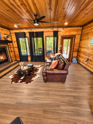 A log cabin interior with a fireplace, leather sofa, and a ceiling fan. - Waterfront: Hot tub, pool table, river, fishing (McCurtain County)