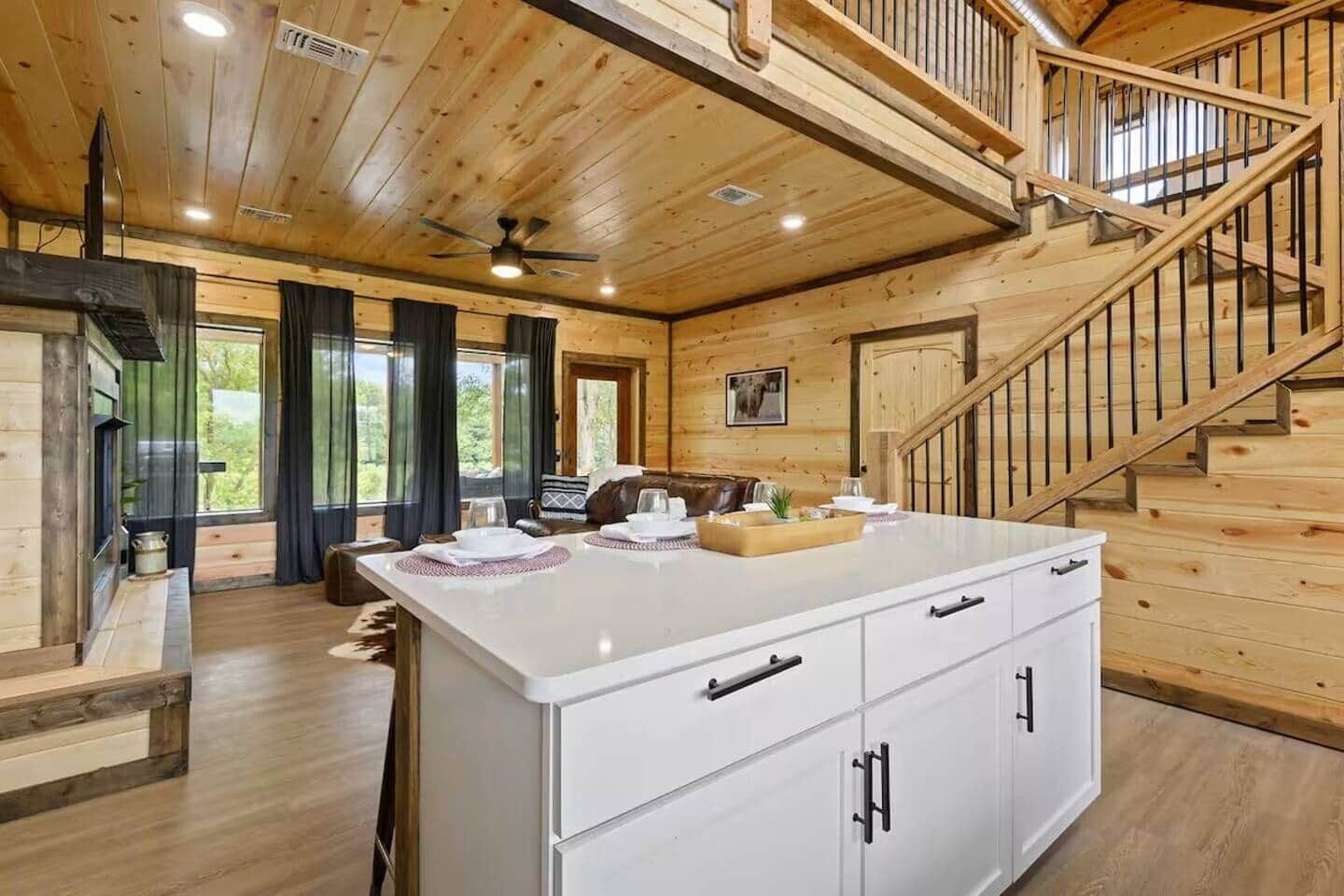 A wooden interior with a central island, white cabinetry, a staircase, and a view through a window.