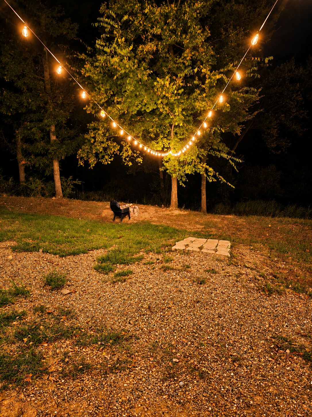 A nighttime outdoor setting with string lights, a fire pit, and a tree.