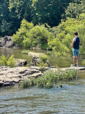 A person standing on rocks by a river with dense greenery in the background. - Waterfront: Hot tub, pool table, river, fishing (McCurtain County)