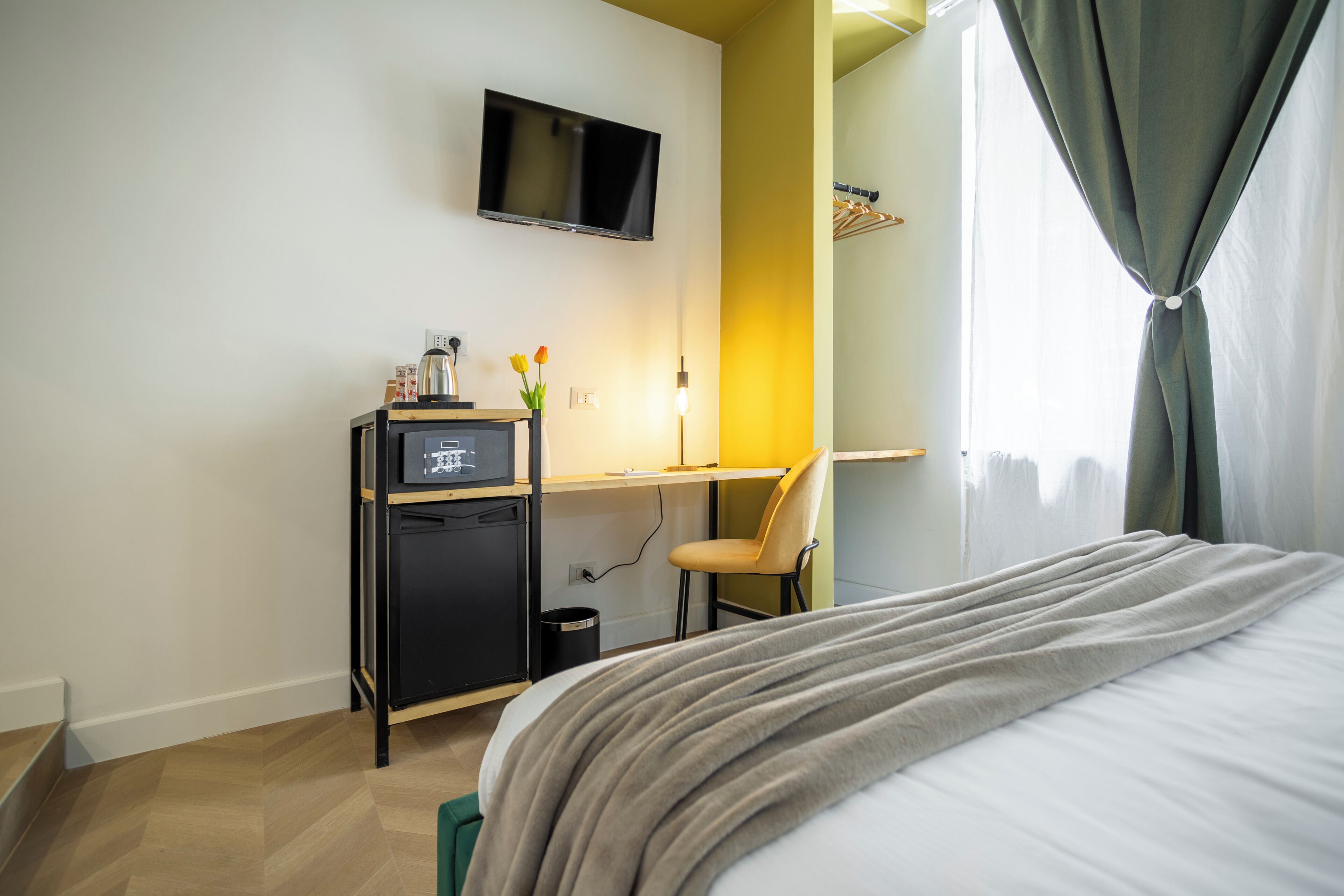 Una habitación de hotel con cama, escritorio con computadora, televisor montado en la pared y ventana con cortinas.