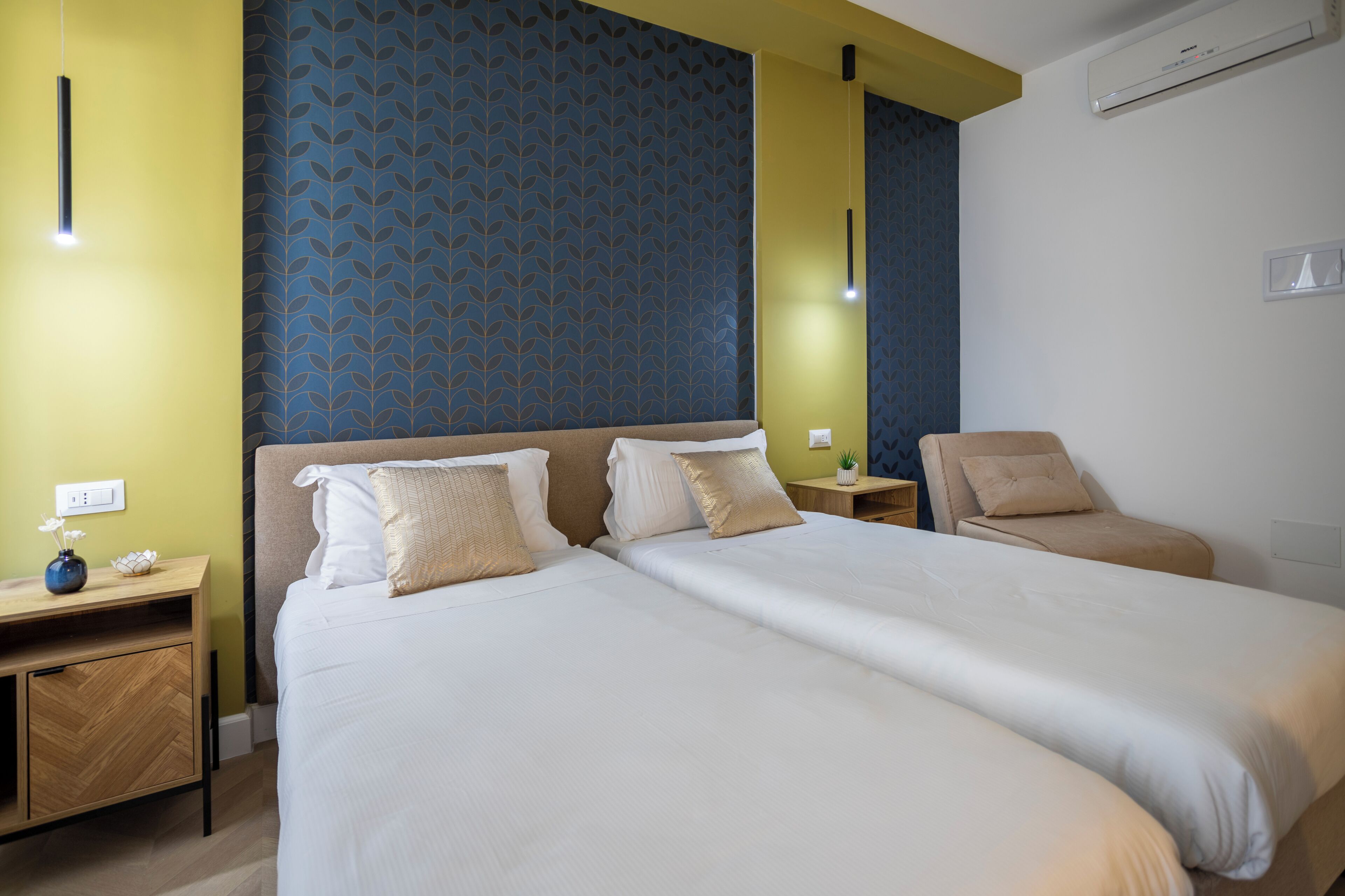 A hotel room with two beds, a bedside table with a vase, a chair, and a patterned wall.