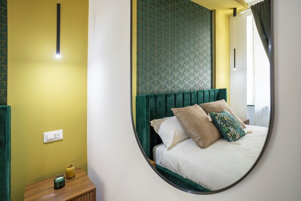 Una cama con cabecera verde y varios almohadones, una mesita de noche con lámpara y un enchufe de pared.