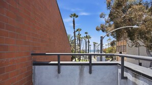 A concrete ledge with a metal railing, a brick wall, and a street with palm trees and a building in the background. - Lovely Beachside 2-Bedroom Apartment (Santa Monica)