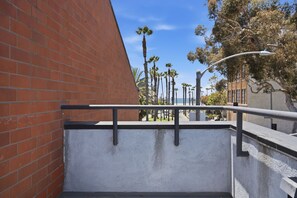 A concrete ledge with a metal railing, a brick wall, and a street with palm trees and a building in the background. - Lovely Beachside 2-Bedroom Apartment (Santa Monica)