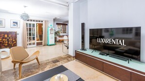 A modern living room with a flat-screen TV, a sofa, a coffee table, and a dining area in the background. - LXR Villa Elysée: 4 Bedrooms, Pool, and Amenities (AN)
