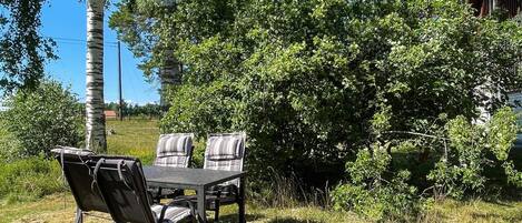 An outdoor seating area with a table and chairs, surrounded by dense foliage and a clear sky.