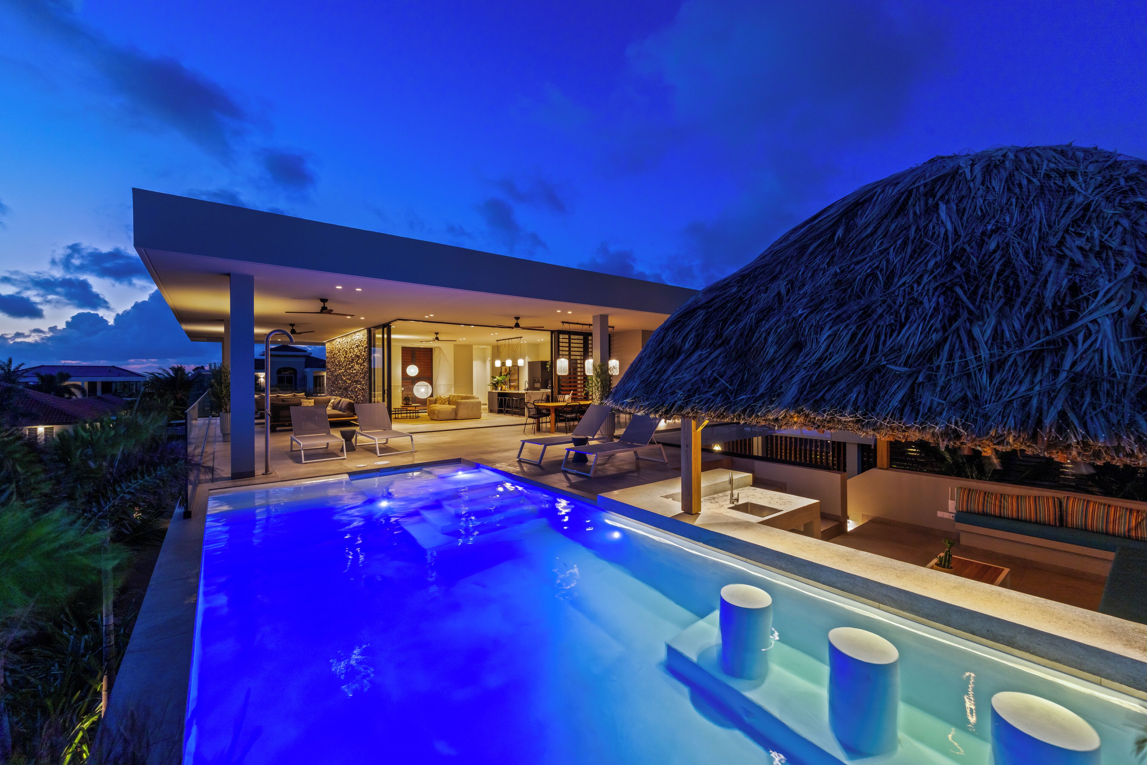 A modern pool area with a thatched roof structure, lounge chairs, and a dining area.