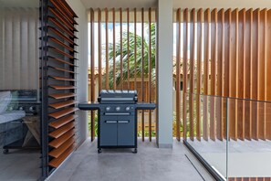 A modern outdoor kitchen area with a portable grill, a seating area with a sofa, and a view of palm trees through vertical wooden slats. - Villa Quatrosol - an exclusive private resort under the sun! (Jan Thiel)