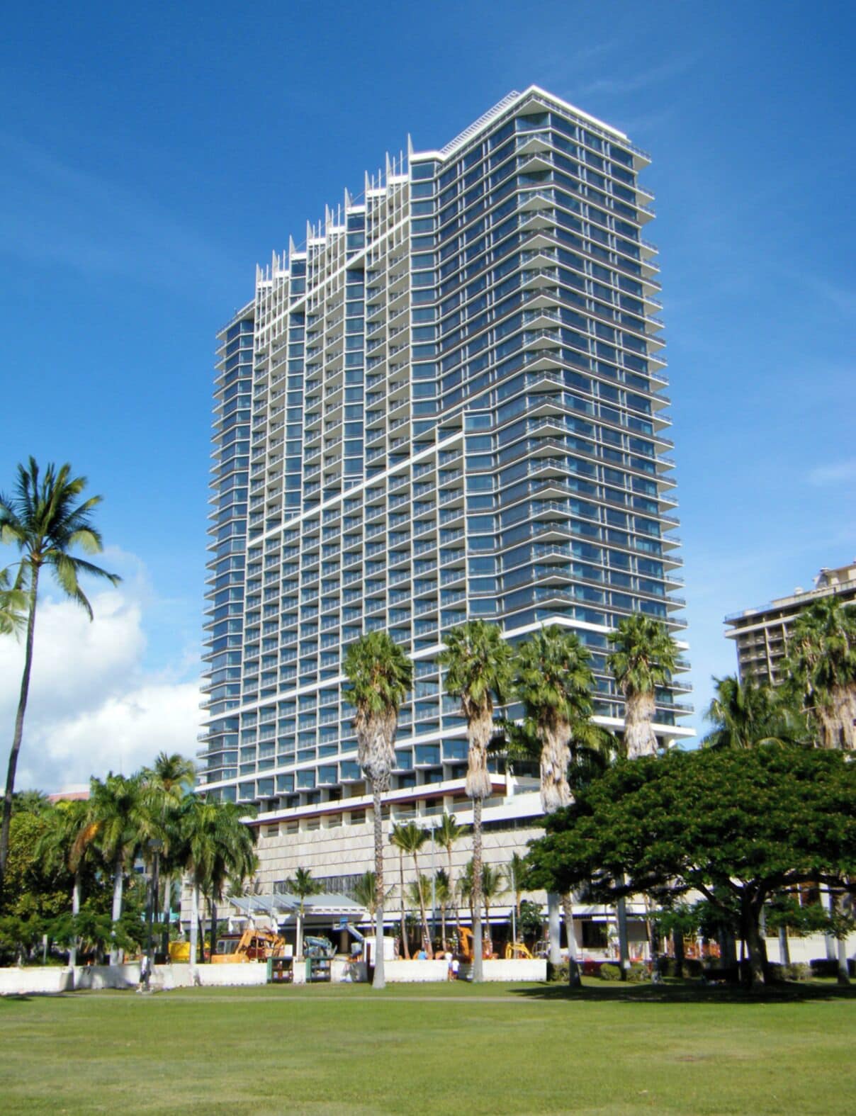 A tall, modern hotel with a grid-like facade, surrounded by palm trees and a grassy area.