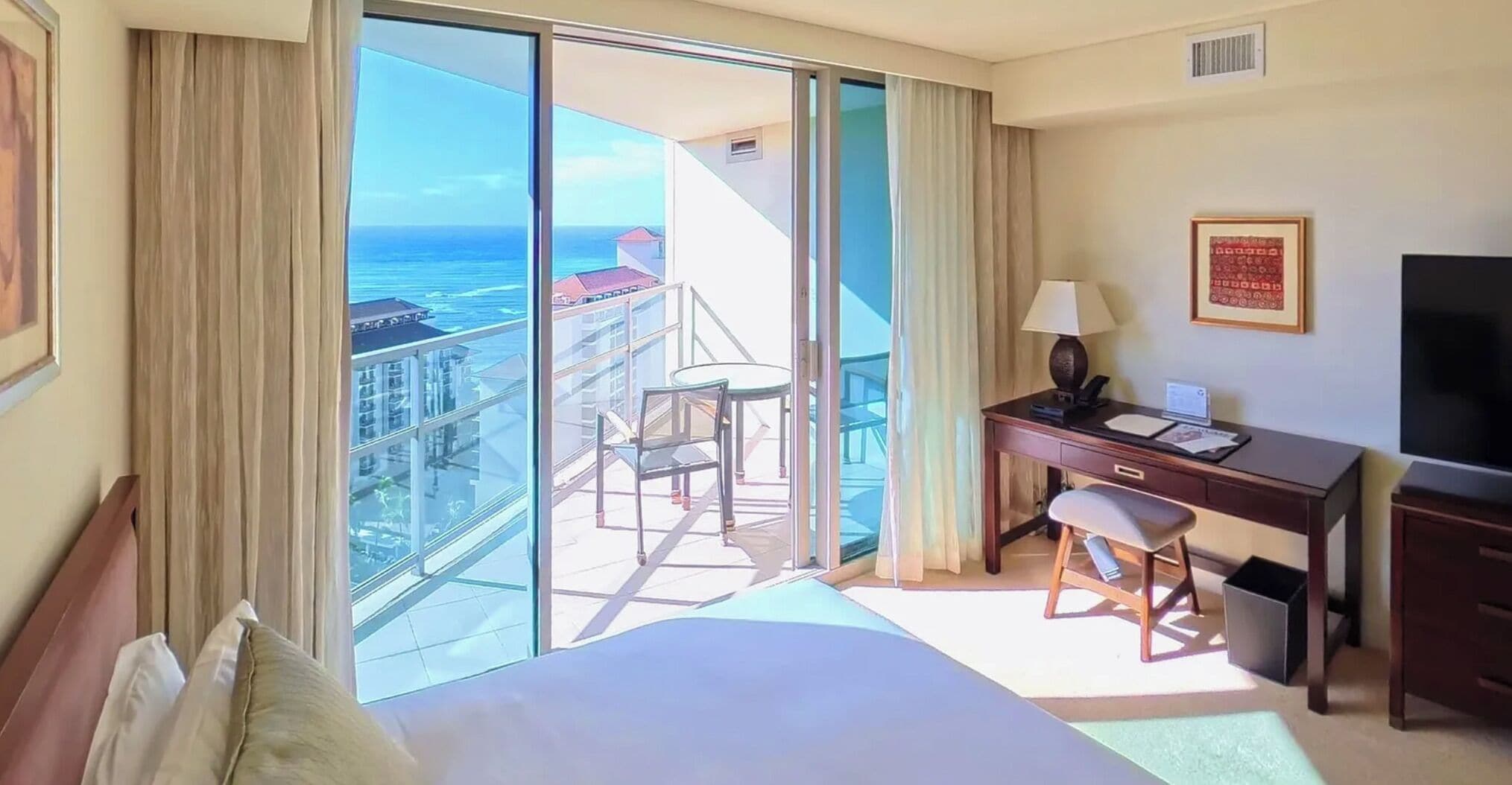 A hotel room with a bed, desk, chair, and a view of the ocean through large windows.