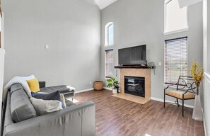Ein Wohnzimmer mit grauem Sofa, Kamin, Fernseher und einem Fenster mit Jalousien.