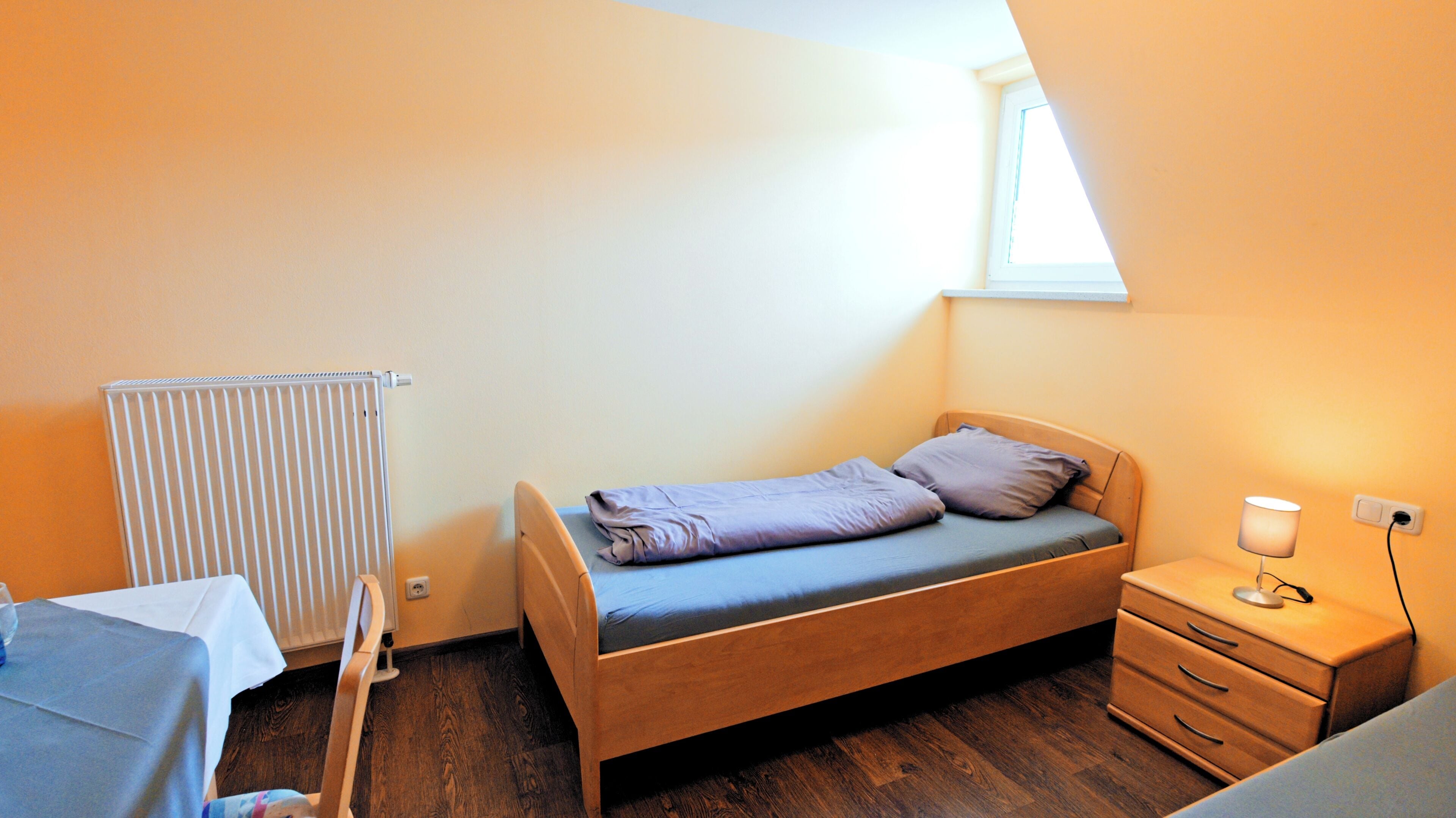 A small, single-bed room with a wooden bed, a bedside table with a lamp, a radiator, and a window.