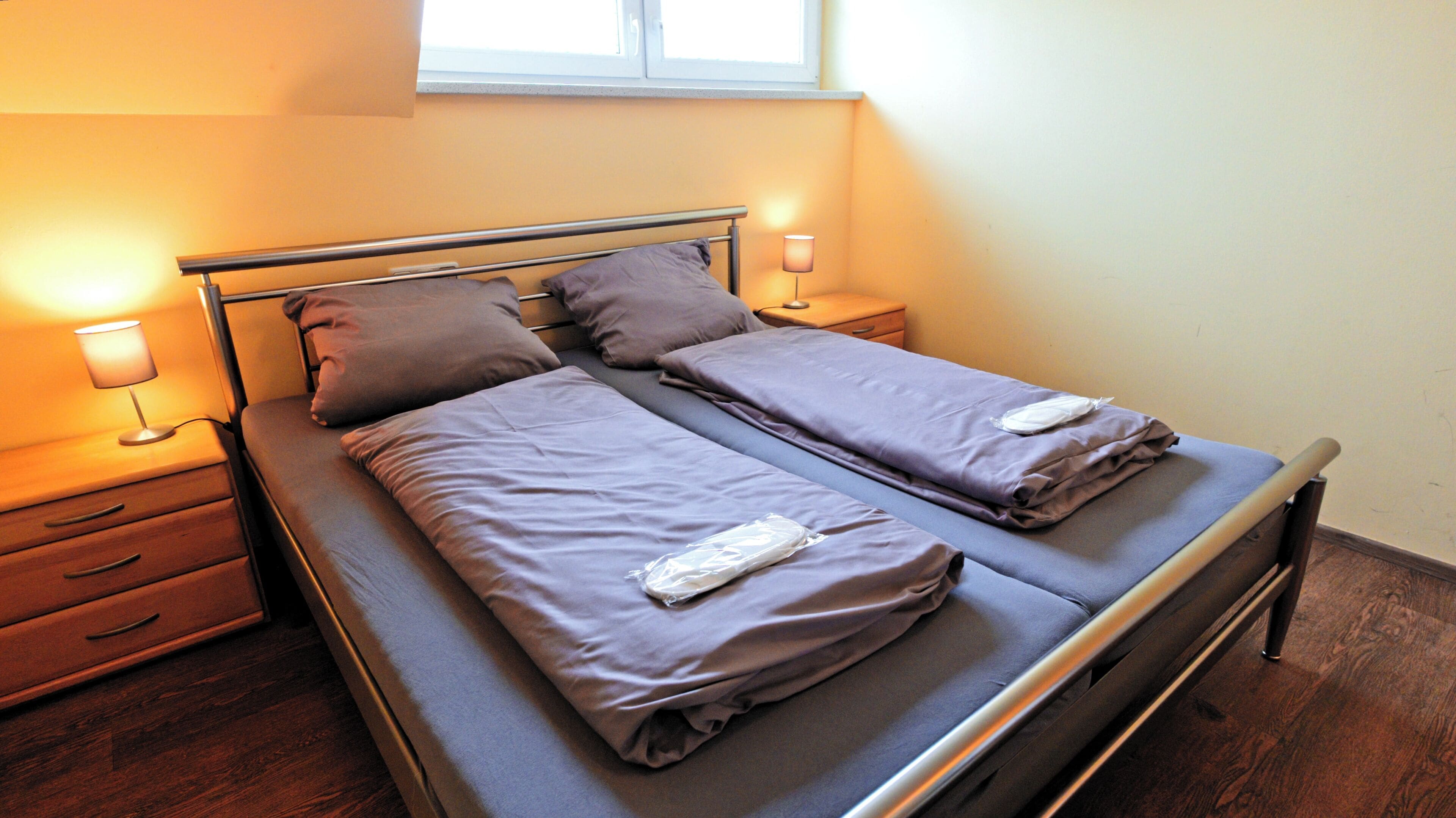 Ein Doppelbett mit zwei Kissen und einer Decke, ein Nachttisch mit einer Lampe sowie ein Fenster über dem Bett.