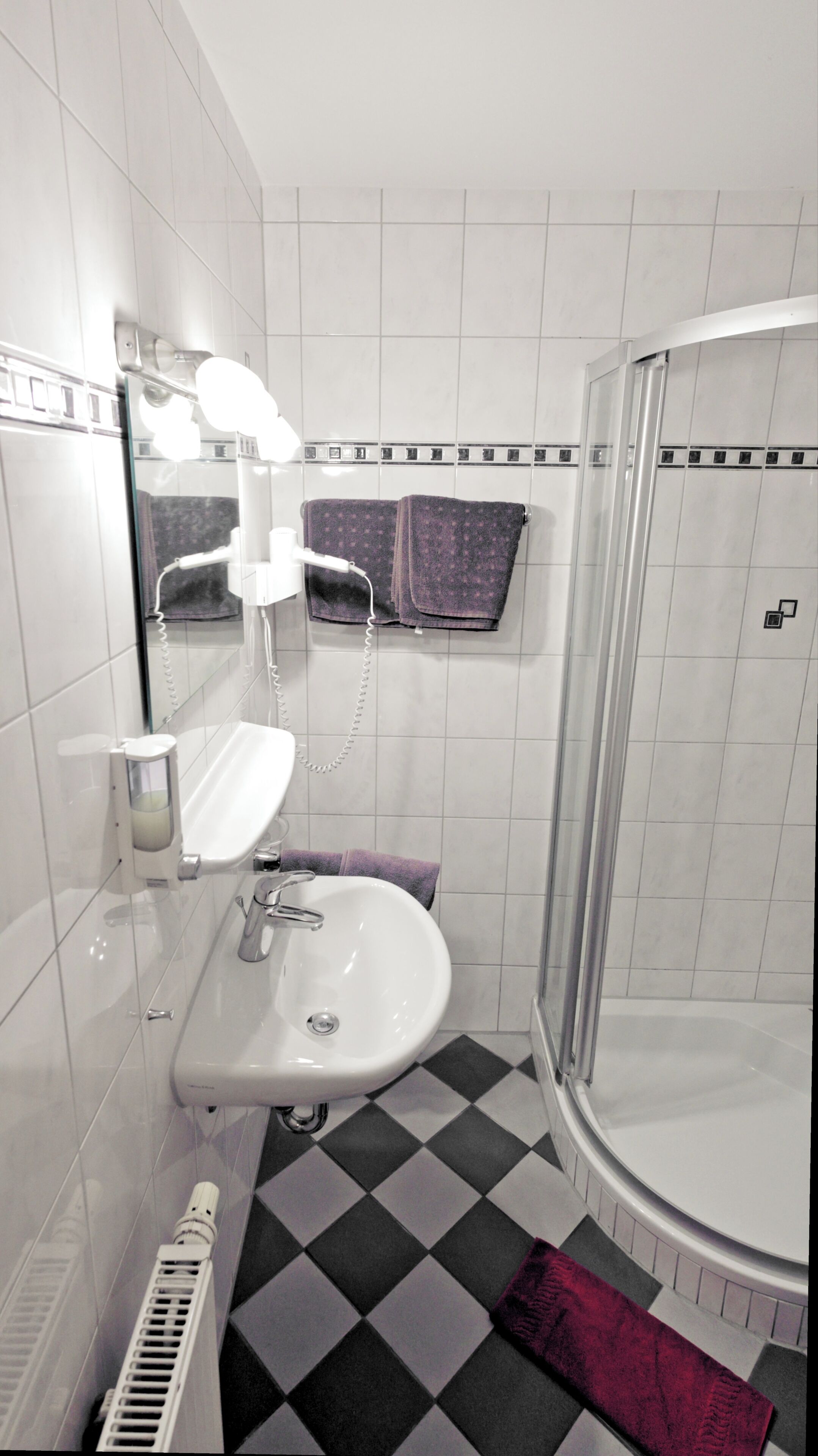 Un baño con un lavamanos blanco, un espejo, un perchero para toallas, una ducha y un piso a cuadros.