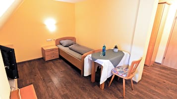 A small room with a bed, a table with a chair, a bedside table, and a wall socket.