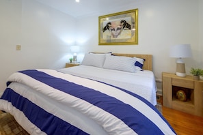 Un dormitorio con una cama grande, una mesita de noche con una lámpara, un cuadro en la pared y una planta pequeña sobre la mesita.