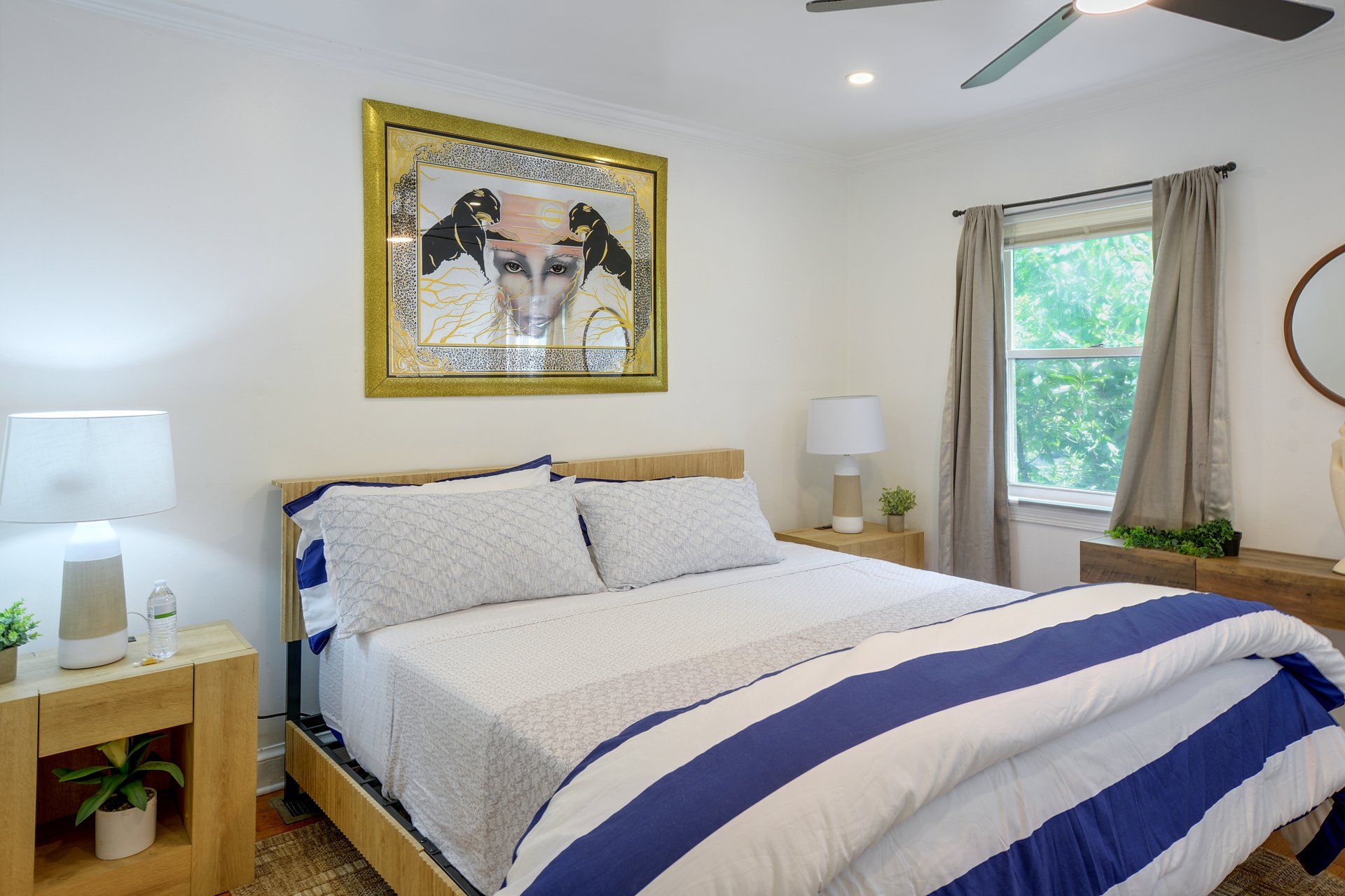 Une chambre à coucher comprenant un lit, deux tables de chevet, un ventilateur de plafond, une fenêtre avec des rideaux et une œuvre d’art accrochée au mur.