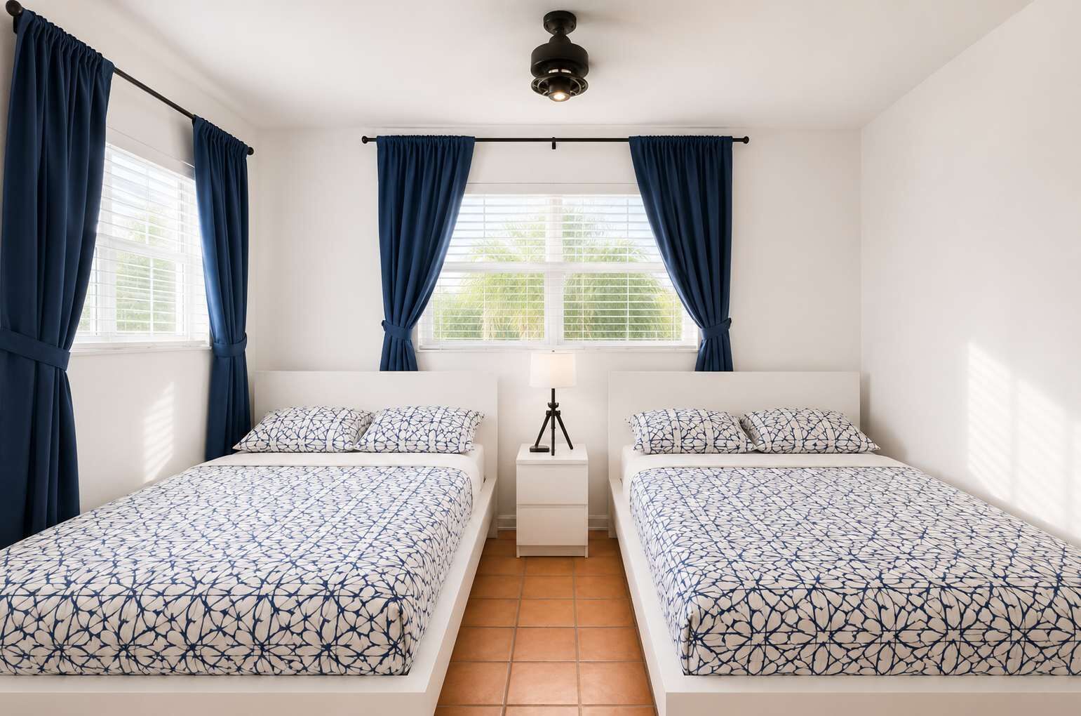 A bedroom with two beds, a window with curtains, a ceiling fan, and a nightstand with a lamp.
