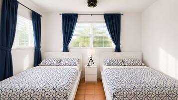 A bedroom with two beds, a window with curtains, a ceiling fan, and a nightstand with a lamp.