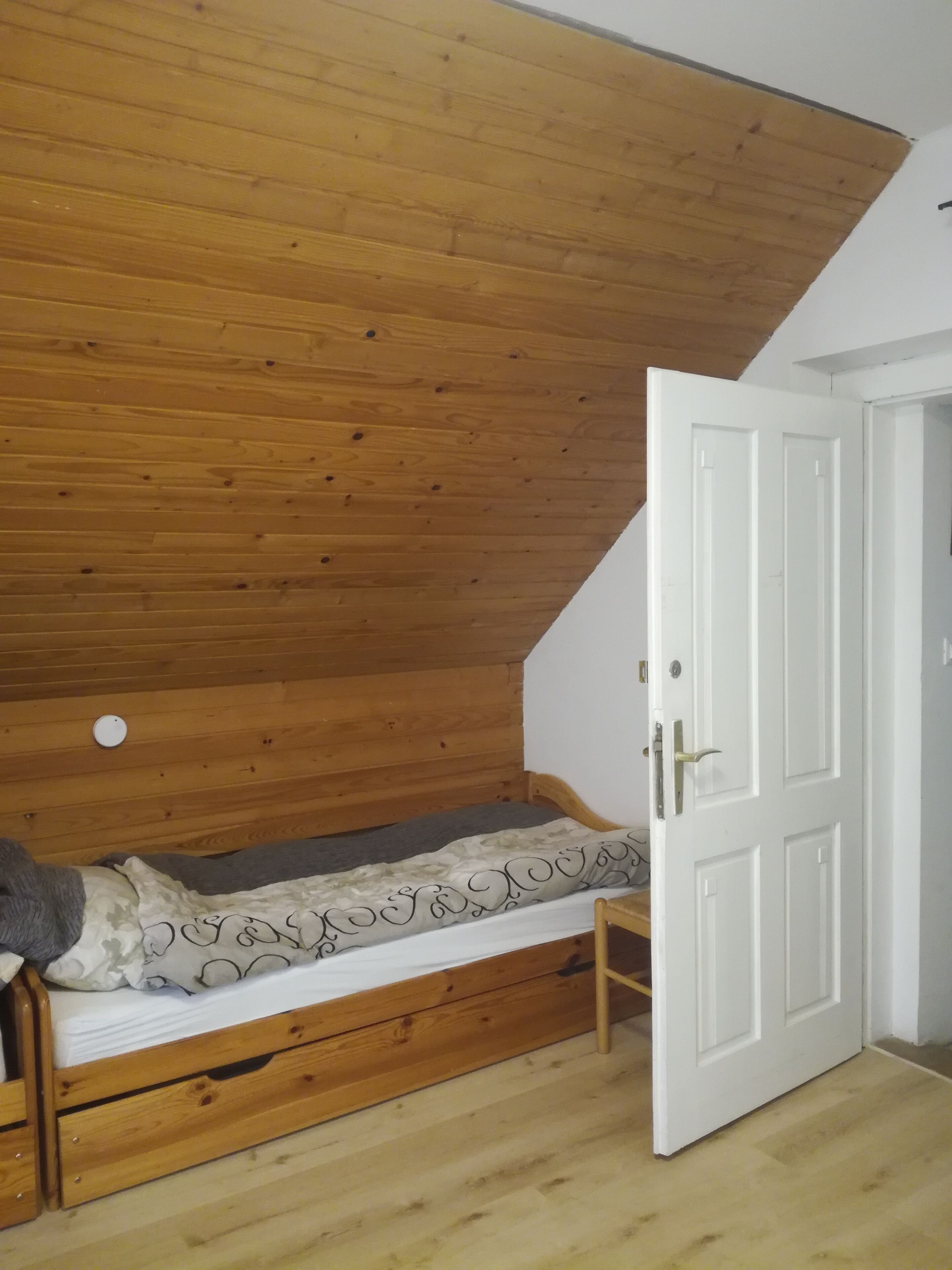 Ein Einzelbett aus Holz in einem Zimmer mit schrägen Wänden und einer weißen Tür.